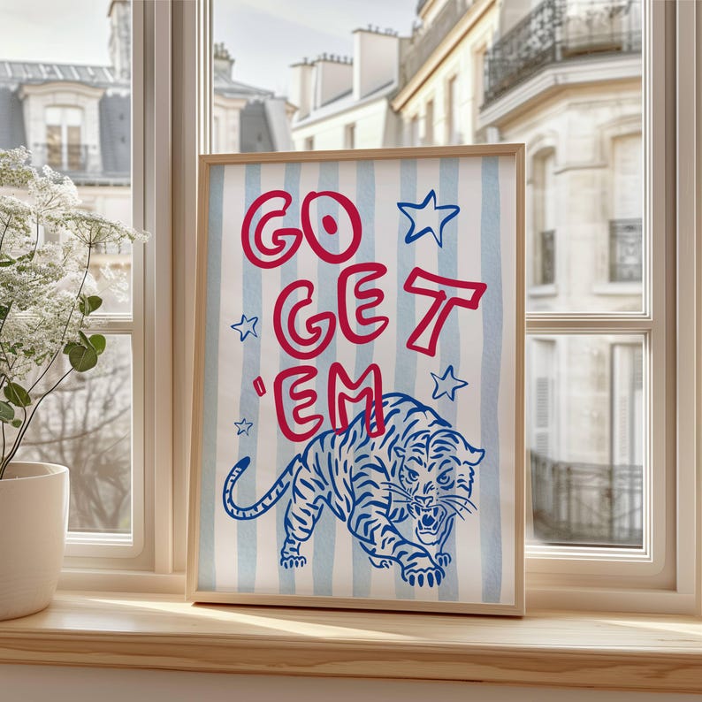 May include: A framed art print with the words "GO GET 'EM" in red, with blue stars and a blue tiger illustration on a striped background. The artwork is displayed on a wooden windowsill.