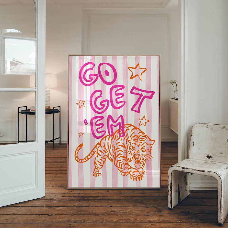 K&ouml;nnte beinhalten: Gerahmter Kunstdruck mit einer Tigerillustration in Orange und Wei&szlig; und dem pinkfarbenen Text "GO GET 'EM". Der Hintergrund hat rosa und wei&szlig;e vertikale Streifen mit Sternakzenten. Das Kunstwerk wird in Innenr&auml;umen ausgestellt.