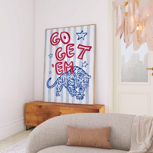 May include: A framed art print featuring a blue and white striped background with the words "GO GET 'EM" in red. A blue tiger illustration is at the bottom. The artwork is displayed above a wooden cabinet, with a light-colored sofa in the foreground.