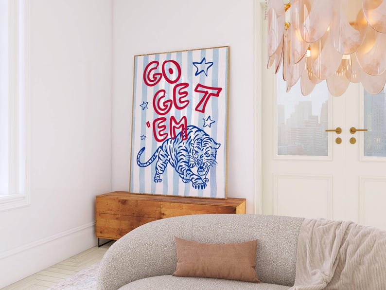 May include: A framed art print featuring a blue and white striped background with the words "GO GET 'EM" in red. A blue tiger illustration is at the bottom. The artwork is displayed above a wooden cabinet, with a light-colored sofa in the foreground.