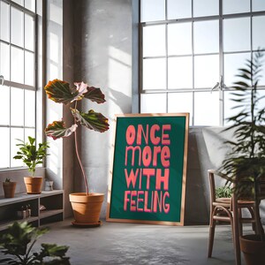 May include: A framed green poster with the text "ONCE more WITH FEELING" in pink letters. The poster is hanging on a wall in a room with plants and a wooden chair.