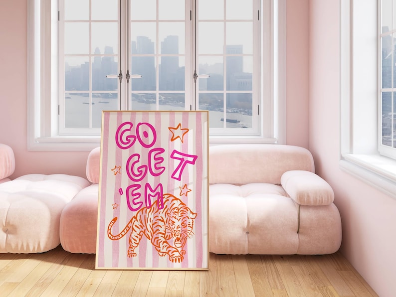 K&ouml;nnte beinhalten: Ein gerahmter Kunstdruck mit den Worten "GO GET 'EM" in Pink und Orange sowie einer Tigerillustration. Das Kunstwerk ist vor einem gestreiften Hintergrund aus Rosa und Wei&szlig; platziert. Der Druck wird in einem Raum mit rosa M&ouml;beln ausgestellt.