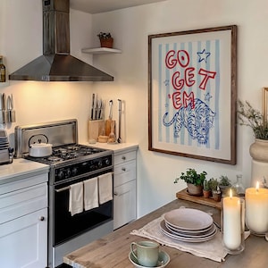 May include: A kitchen scene featuring a framed artwork with the text "GO GET 'EM" and a blue tiger illustration. The kitchen includes a stainless steel range hood, stove, and a wooden table set with plates and a teacup. Two lit candles add warmth.