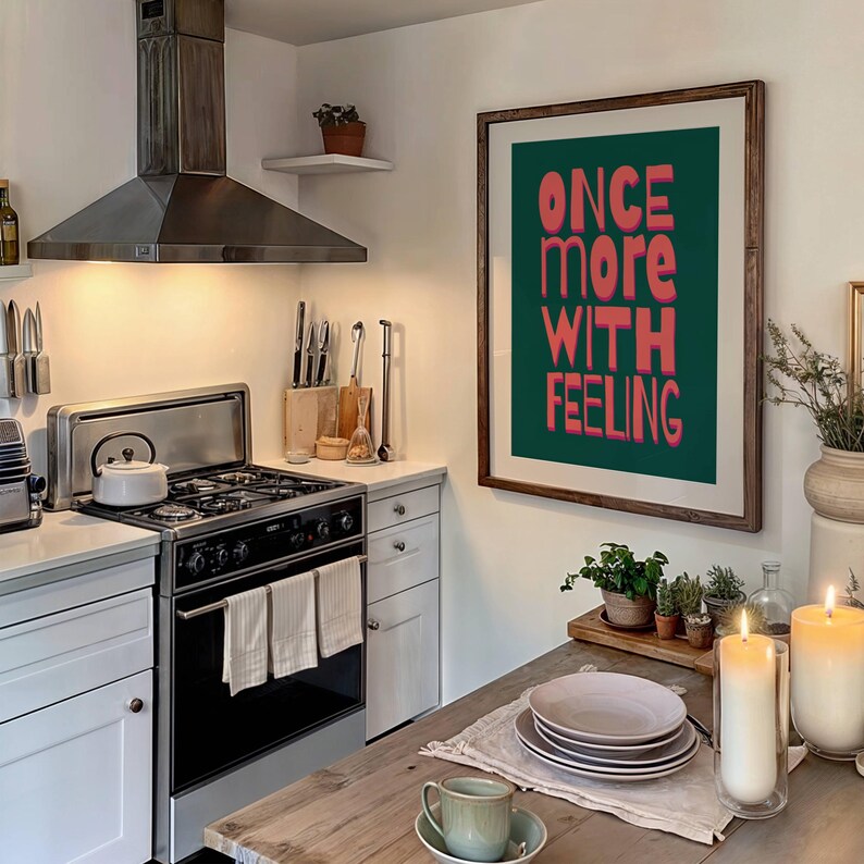 May include: A framed print with the text "ONCE MORE WITH FEELING" in pink letters on a dark green background. The print is hanging on a white wall in a kitchen.