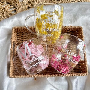 Peut inclure: Trois tasses en verre à double paroi avec anse. Chaque tasse contient des fleurs séchées jaunes, roses ou magenta. Les tasses présentent des inscriptions blanches incluant des prénoms comme Anaëlle, Meilleure Marraine et Matheo.