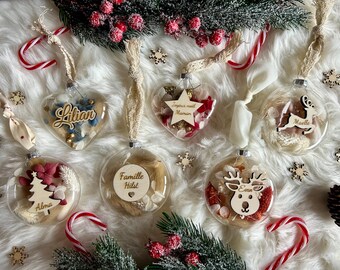 Peut inclure: Plusieurs boules de Noël transparentes en verre remplies de fleurs séchées et d'éléments décoratifs. Certaines sont en forme de cœur, d'autres rondes, et comportent des découpes en bois avec des noms et des motifs festifs. Des cannes en sucre et des branches de pin entourent les boules.