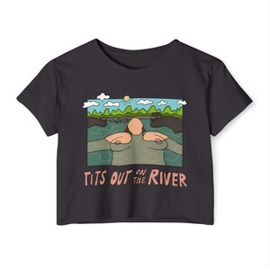 May include: Black crop top with a cartoon graphic of a person in a river with the text "Tits Out on the River". The graphic features a sunny sky, green hills, and blue water.