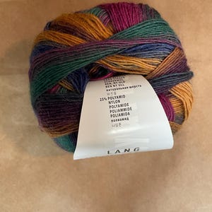 May include: A ball of multicolored yarn with shades of green, purple, orange, and blue. A white label with text in multiple languages is attached to the yarn. The yarn is made of 25% Polyamid and Nylon.