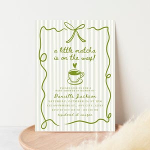 Puede incluir: Invitación a un baby shower con un fondo a rayas verdes y blancas. La invitación presenta texto e ilustraciones verdes, incluyendo una taza de té y la frase "a little matcha is on the way!". Los detalles incluyen el nombre de la homenajeada, la fecha, la hora y la información RSVP.