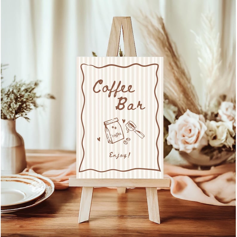 May include: A small wooden easel holds a sign that reads "Coffee Bar" in brown script. The sign has a striped background and a brown border. Below the text is a coffee-themed illustration. The sign is on a wooden table.