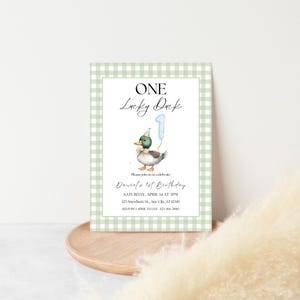 May include: A green and white gingham patterned invitation card with a cartoon duck wearing a party hat and holding a blue number one balloon. The text on the card reads "One Lucky Duck" and includes details for a first birthday party.