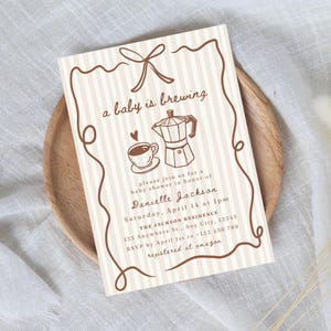May include: A baby shower invitation with a coffee theme. The invitation features a brown and white striped background with a decorative border. The text "a baby is brewing" is at the top, along with a coffee cup and a moka pot illustration.