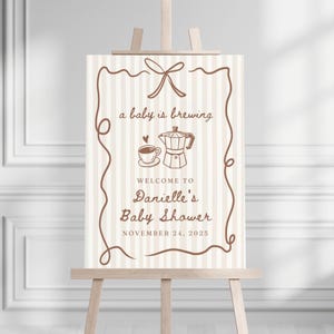 May include: A beige and brown baby shower sign with a coffee pot and cup illustration. The text reads "a baby is brewing" and "Welcome to Danielle's Baby Shower November 24, 2025".