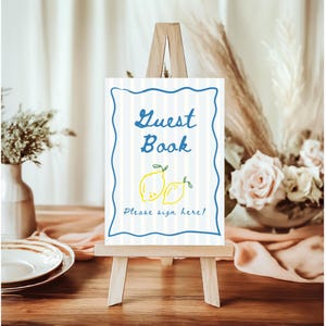 K&ouml;nnte beinhalten: Eine Holzstaffelei zeigt ein "G&auml;stebuch"-Schild mit blau-wei&szlig; gestreiftem Hintergrund. Das Schild hat einen blauen, wellenf&ouml;rmigen Rand, den Schriftzug "Guest Book" in Blau und Illustrationen von zwei gelben Zitronen. Der Text "Please sign here!" steht unten.