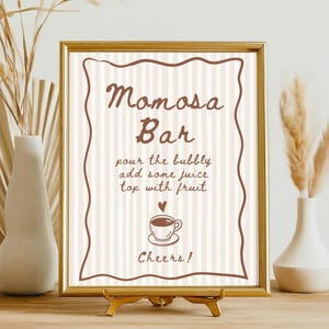 May include: A framed sign with the words "Momosa Bar" in brown script. The sign includes instructions and a coffee cup graphic. The frame is gold, and the sign is displayed on a gold stand.