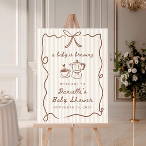 May include: A beige and brown welcome sign with a coffee pot and cup illustration. The text reads "a baby is brewing" and "Welcome to Danielle's Baby Shower November 24, 2025".