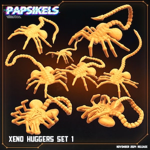 May include: A collection of light orange Xenomorph facehugger figures. The figures have multiple legs and long, segmented tails. The text "Papsikels" is at the top, and "Xeno Huggers Set 1" is at the bottom. The release date is November 2024.