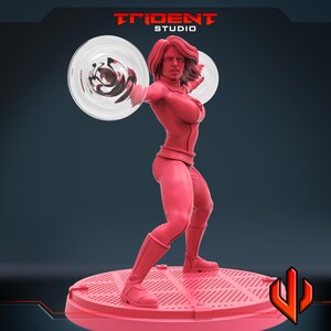 May include: A red figurine of a female superhero in a dynamic pose, holding two transparent energy discs. The figure is wearing a form-fitting suit and boots. The base is a circular platform. The Trident Studio logo is visible.