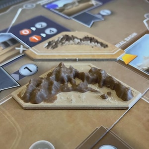 May include: A board game with brown mountain terrain pieces. The game board features various game elements, including number markers and text labels. The mountain pieces are tan with brown terrain details.