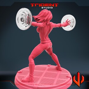 May include: A pink figurine of a female superhero in a dynamic pose, holding two transparent energy discs. The figure is on a pink circular base with a perforated design. The Trident Studio logo is visible at the top.