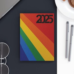 May include: A rainbow-coloured 2025 planner with a black laptop, two pens, a pair of glasses, and a plate with toast and a sausage patty on a white surface.