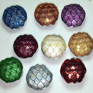 May include: Ten round, decorative, silver-toned compacts with a variety of glitter finishes. The compacts feature a dragon scale design in colors such as blue, green, red, gold, silver, and purple.