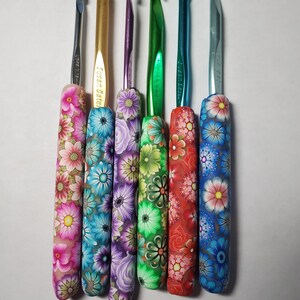 May include: Six crochet hooks arranged vertically, each featuring a unique polymer clay handle decorated with vibrant floral patterns. The hooks vary in metallic colors including silver, gold, purple, green, teal, and blue, with floral designs.