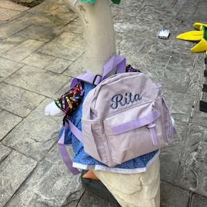 以下が含まれることがあります： 青いドレス、カラフルなジャケット、そして「Rita」と刺繍された薄紫色のバックパックを身に着けた白いガチョウの像。ガチョウは頭に緑のリボンをつけており、屋外に立っています。