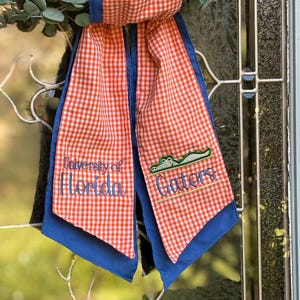 May include: A decorative orange and white gingham ribbon with blue trim. The ribbon features the text "University of Florida" and "Gators" embroidered in blue, along with a green alligator graphic.