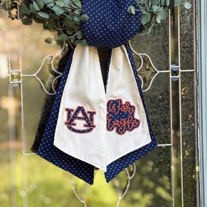 May include: A navy blue and white polka dot graduation sash with the words "War Eagle" and the letter "A" in the center. The sash is hanging on a glass door with a green wreath in the background.