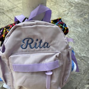 以下が含まれることがあります： 青い筆記体で「Rita」と刺繍された、小さなラベンダー色のバックパック。 ラベンダー色のストラップと留め金が付いたフロントポケットがあります。 追加のラベンダー色のストラップとアクセントが見えます。