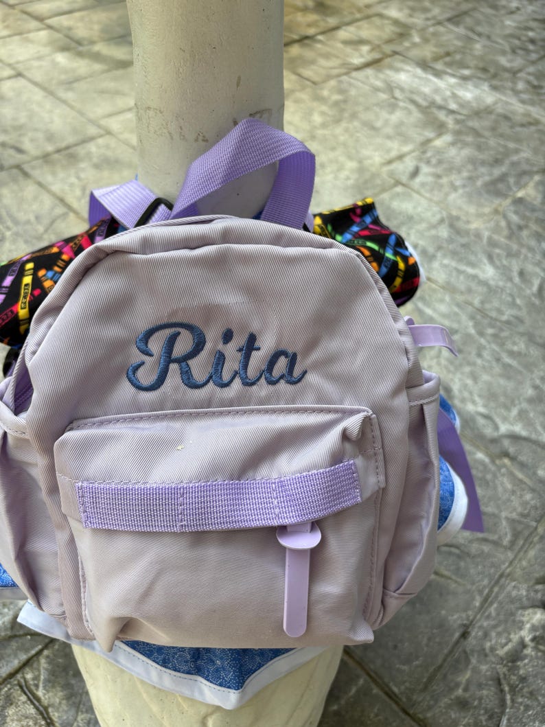 以下が含まれることがあります： 青い筆記体で「Rita」と刺繍された、小さなラベンダー色のバックパック。 ラベンダー色のストラップと留め金が付いたフロントポケットがあります。 追加のラベンダー色のストラップとアクセントが見えます。