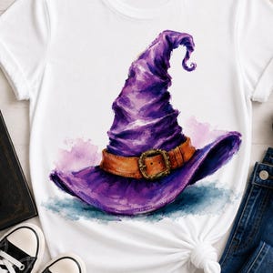 May include: A white t-shirt featuring a large, watercolor-style graphic of a purple witch hat. The hat has a prominent, textured brown leather-look band with a gold-toned buckle around the base. The design displays deep purple shading.