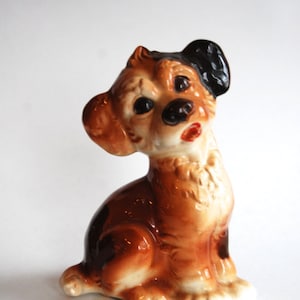 May include: A ceramic figurine of a brown and black dog with a white chest and paws. The dog is sitting with its head tilted to the side and its mouth slightly open.