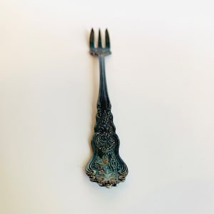 May include: A silver fork with an ornate, detailed handle. The fork has a tarnished finish and is designed for serving desserts or appetizers.