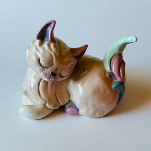 May include: A ceramic cat figurine in a resting pose, with closed eyes and a serene expression. The cat is primarily peach-colored, with accents of pink, purple, and green on the ears, tail, and paws. Delicate black details enhance the facial features.