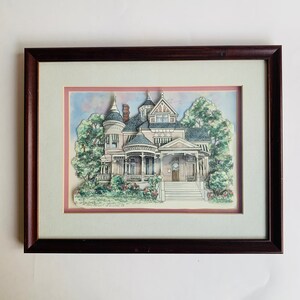 May include: A framed illustration of a pink Victorian house with a white picket fence and a lush green lawn. The house has a large porch with white columns and a white door. The illustration is surrounded by a brown frame.