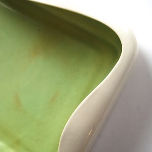 Puede incluir: Un plato de cerámica blanca con un interior esmaltado verde. El plato tiene una forma única y ondulada, y es perfecto para servir aperitivos o guardar objetos pequeños.