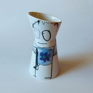 May include: A white ceramic vase with a whimsical, hand-drawn design. The vase features a cartoon-like face with large eyes and a blue shirt with a purple flower. The vase has a unique, angled opening.