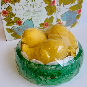 Puede incluir: Un recipiente de pl&aacute;stico verde con una tapa transparente, que contiene tres jabones amarillos en forma de p&aacute;jaro. El recipiente tiene un dise&ntilde;o floral y el texto "AVON LOVE NEST SOAP DISH and THREE "SPECIAL OCCASION" FRAGRANCED SOAPS NET WT 1.5 OZ EACH TOTAL NET WT 4.5 OZ" est&aacute; impreso en la caja.