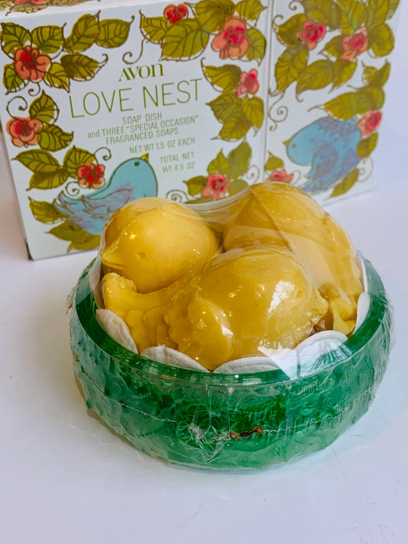 Puede incluir: Un recipiente de pl&aacute;stico verde con una tapa transparente, que contiene tres jabones amarillos en forma de p&aacute;jaro. El recipiente tiene un dise&ntilde;o floral y el texto "AVON LOVE NEST SOAP DISH and THREE "SPECIAL OCCASION" FRAGRANCED SOAPS NET WT 1.5 OZ EACH TOTAL NET WT 4.5 OZ" est&aacute; impreso en la caja.