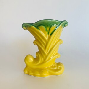 May include: A yellow ceramic vase with a green rim and a stylized design resembling a corn husk. The vase has a unique shape with a curved base and a flared opening.