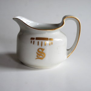 May include: White ceramic creamer with a gold rim and handle. The creamer features gold decorative elements and a stylized gold letter "S" on the front. The creamer is designed for serving cream or milk.
