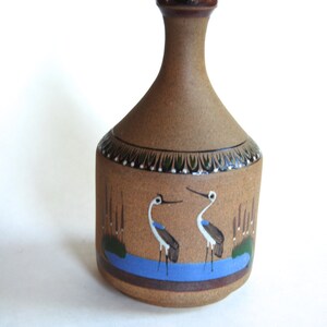 May include: A brown ceramic vase with a narrow neck and a wide base. The vase is decorated with a blue and green scene of two white birds standing in a pond with reeds.