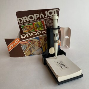 May include: A white and black pen with a black base and a notepad. The pen is attached to the base with a magnet. The base has a sticky pad on the bottom so it can be attached to a phone. The packaging for the product is in the background. The packaging is brown with white text that says "Drop'n Jot Self-Stick Telephone Dial Pen & Note".