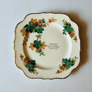 May include: Square, off-white plate with a scalloped gold rim. The plate features a floral design with green leaves and orange flowers. Text on the plate reads "COMPLIMENTS OF ZULAUF & SON GENERAL MERCHANDISE HOME KILLED MEATS ARENZVILLE, ILL."