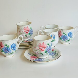 May include: A set of six teacups and saucers with a floral pattern featuring pink, blue, and white sweet pea flowers. The cups and saucers are white with a gold trim.