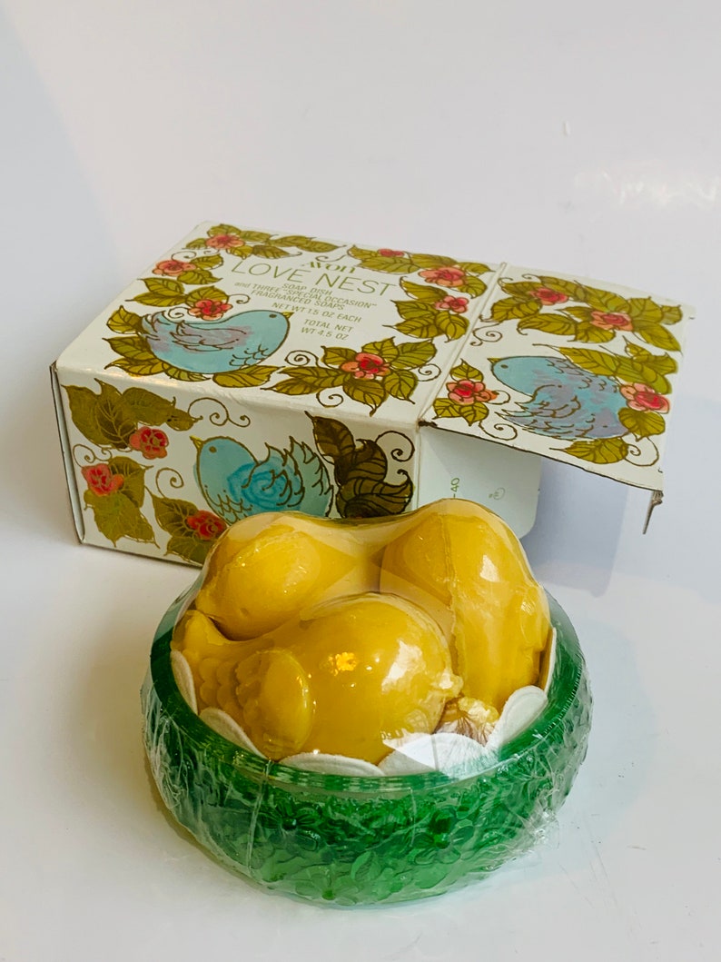 Puede incluir: Un juego de tres jabones amarillos con forma de p&aacute;jaro en un recipiente verde con forma de nido. La caja es blanca con un dise&ntilde;o floral y el texto "Avon Love Nest Soap Dish Three Fragranced Soaps Net Wt 1.5 oz Each Total Net Wt 4.5 oz".