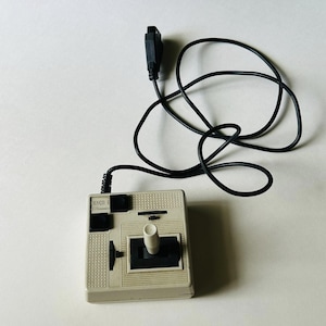 May include: A vintage beige joystick controller with a black cable. The controller has a white joystick, black buttons, and the text "MACH III" printed on the front. The black cable is coiled.