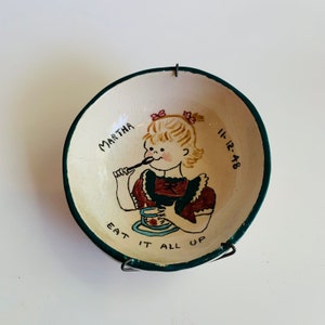 May include: A white ceramic bowl with a green rim features a hand-painted illustration of a girl with blonde hair eating from a bowl. The bowl has the text "Martha 11-12-48 Eat It All Up" painted on it.